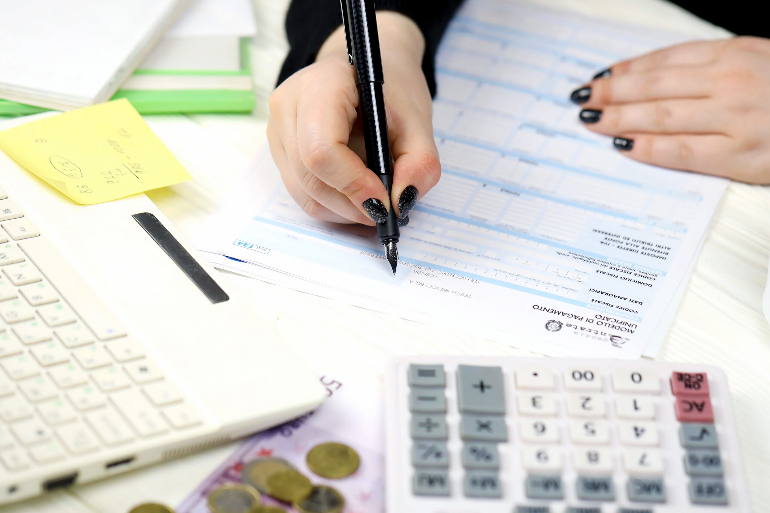 Modern,Country,House,With,The,Large,Lawn,And,A,Wooden mani di donna intente a compilare documenti IMU su una scrivania dove si trovano anche un computer e una calcolatrice.